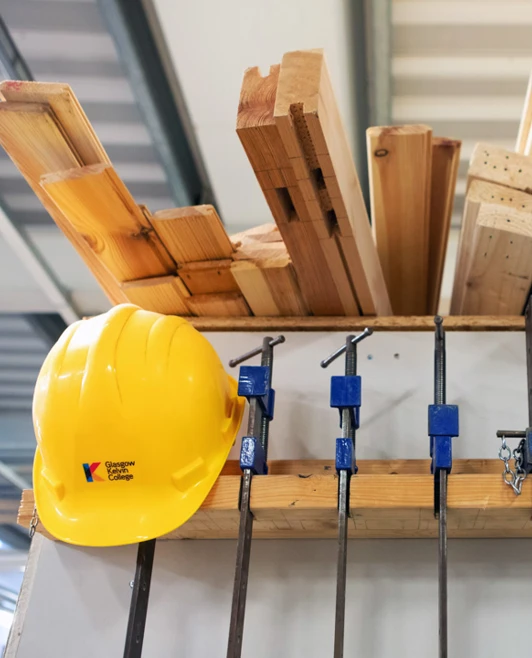 Woodworking equipment including clamps, timber planks, and a bright yellow safety helmet neatly arranged on a storage rack in a joinery workshop. Woodworking equipment including clamps, timber planks, and a bright yellow safety helmet neatly arranged on a storage rack in a joinery workshop.